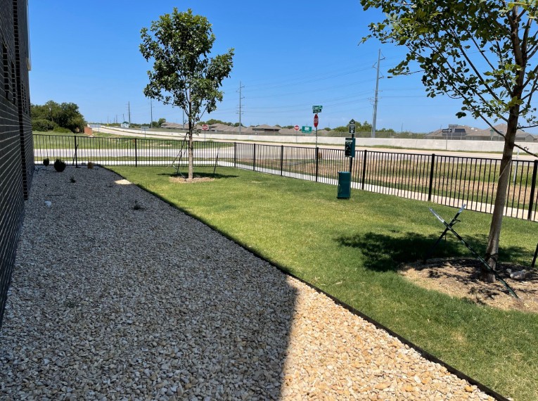 A gravel area is surrounded by a fence and a grassy area with a tree.